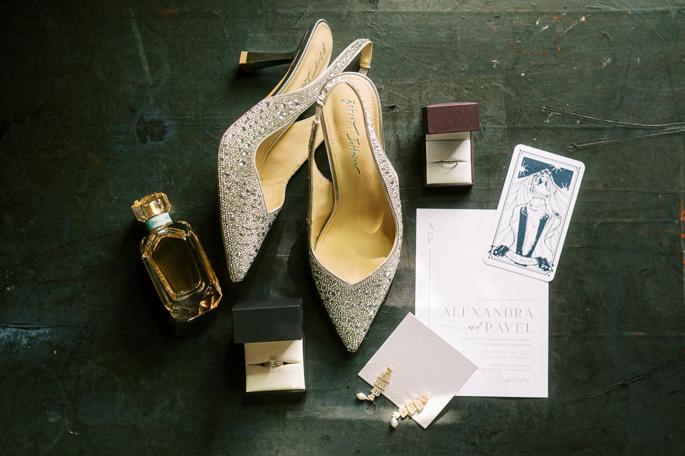 Elegant wedding flat lay: sparkly silver heels, a bottle of perfume, wedding invitations, a ring box with a diamond ring, and statement earrings displayed on a dark textured surface. Perfect for bridal accessory inspiration.