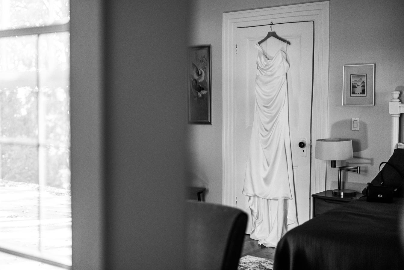 Elegant white wedding dress hanging on a door in a softly lit bedroom, featuring tasteful decor including framed art and a bedside lamp.