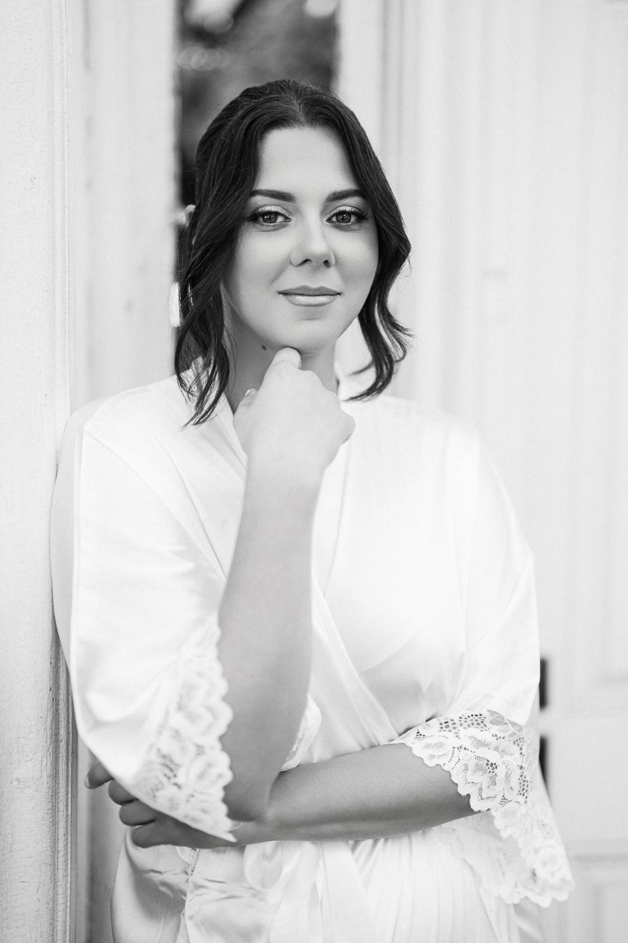Black and white portrait of a woman in a white lace-trimmed robe, thoughtfully posing with her hand under her chin.