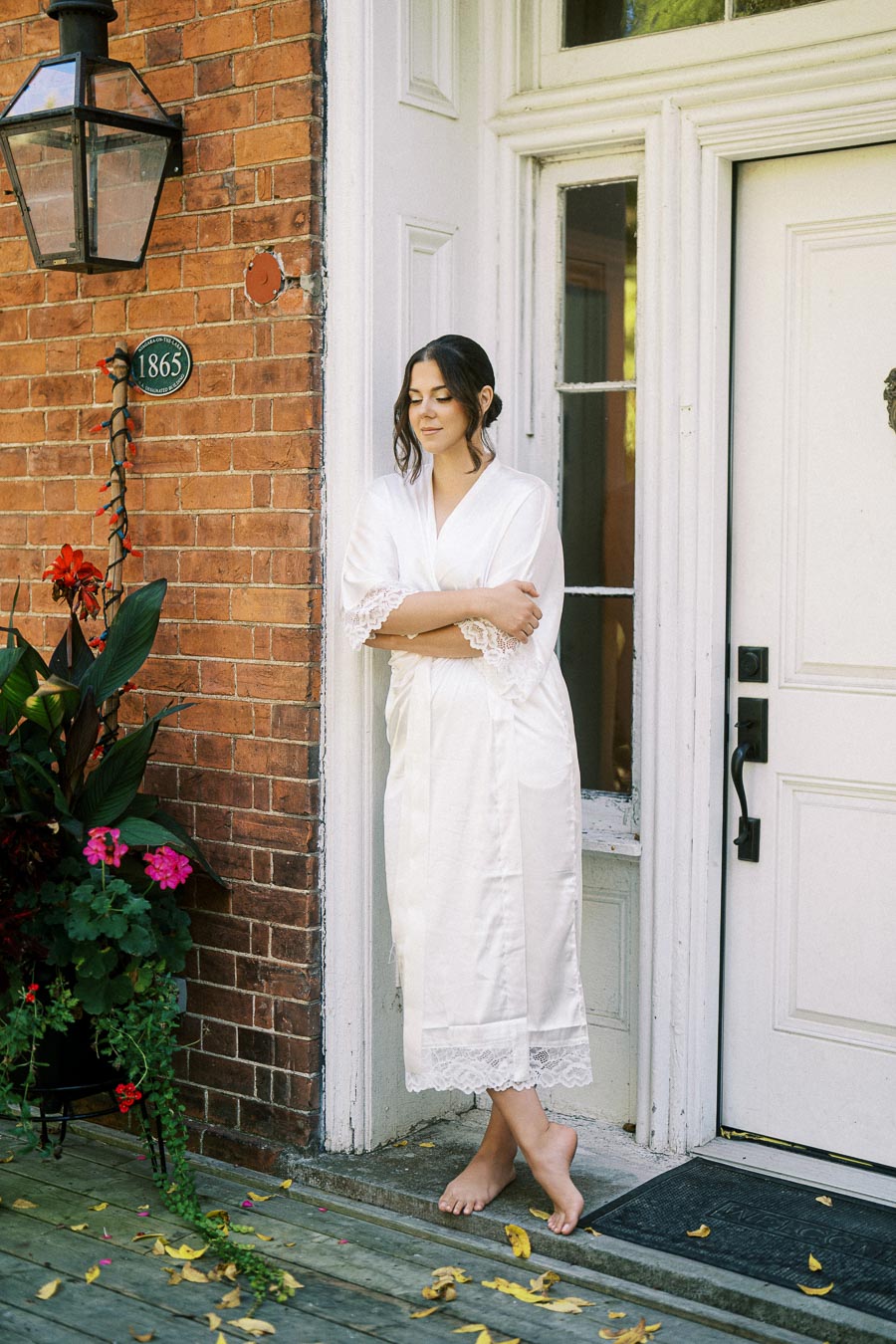 Woman in white robe standing barefoot on a wooden deck, leaning against a brick wall with colorful flowers, in front of a vintage door with a glass lantern above.
