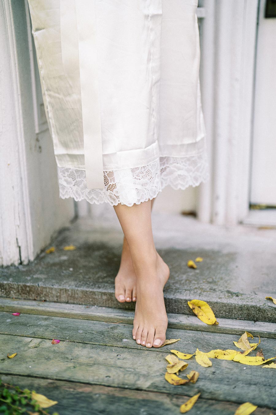 Barefoot person standing on wooden steps with scattered yellow leaves, wearing a long white lace-trimmed garment.