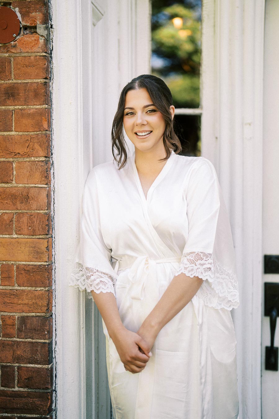 A person wearing a white robe with lace details, smiling while standing against a brick wall next to a door with greenery in the background.