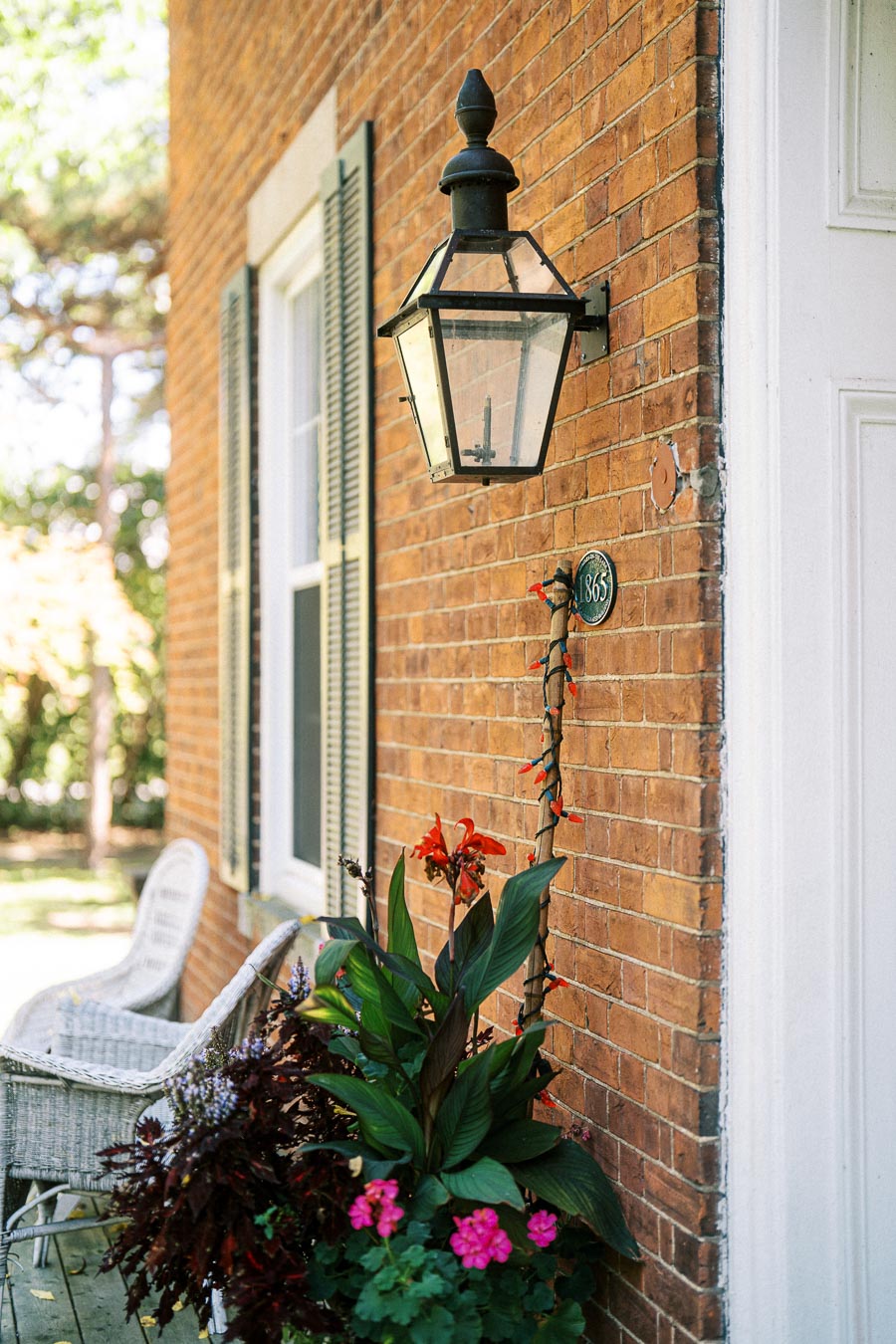 Charming brick house exterior with an antique lantern, window with green shutters, and vibrant flower arrangement in a planter on the porch.
