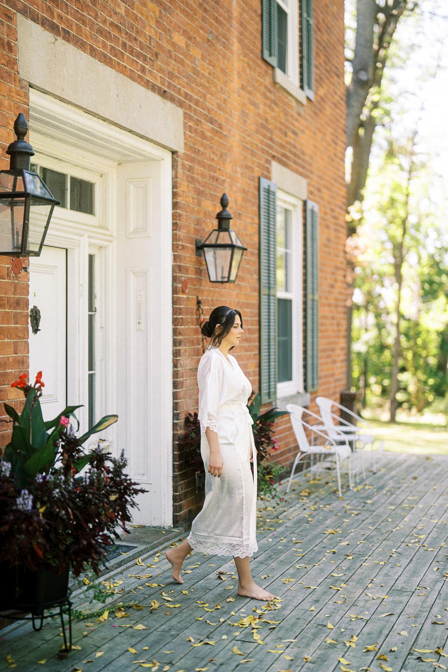 A woman in a white robe walks on a wooden deck beside a brick house with white trim and decorative lanterns, surrounded by potted plants and autumn leaves.