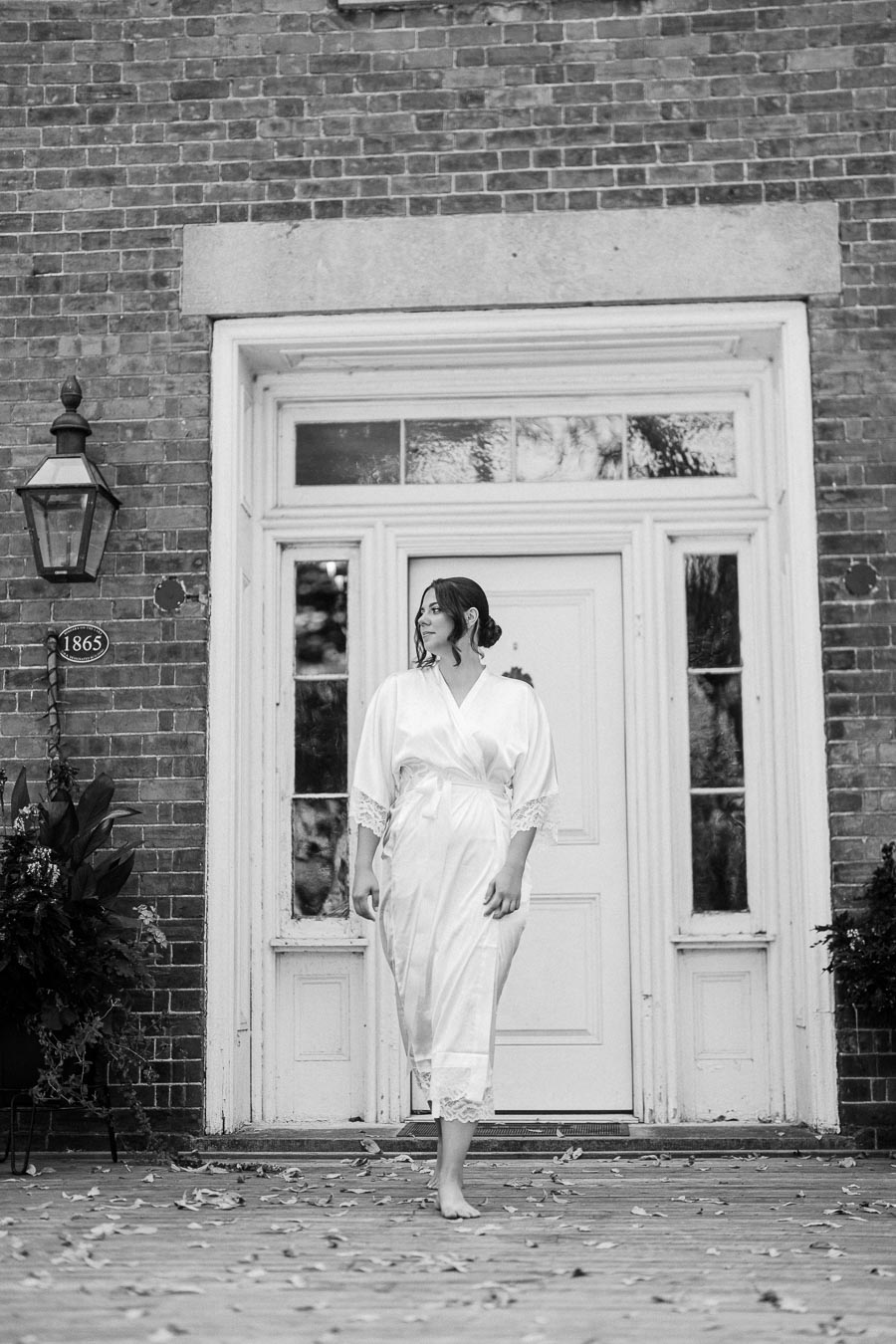 Black and white photo of a woman in a white robe standing outside a brick building with large windows and a vintage lamp.