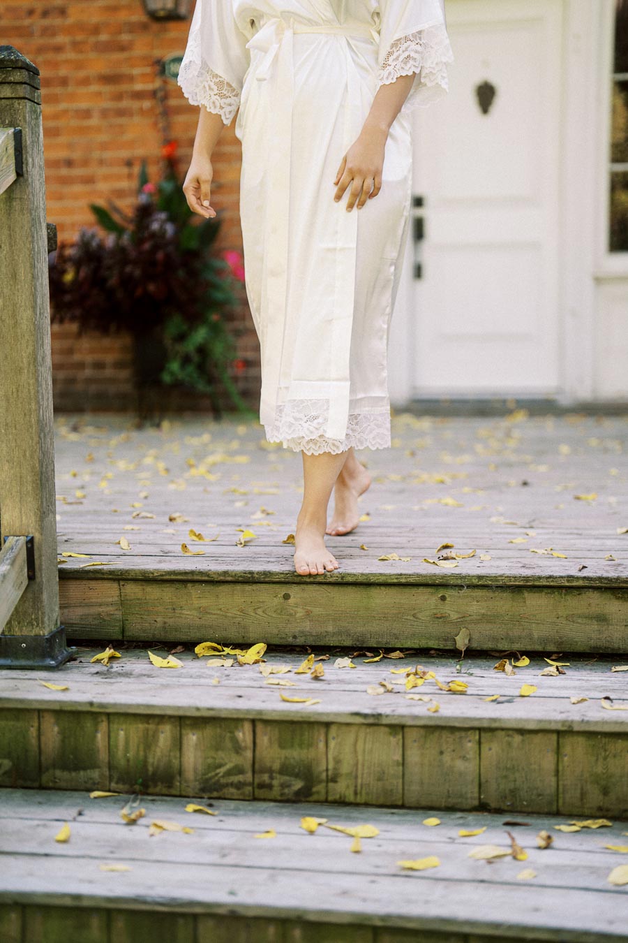 A person in a white lace-trimmed robe walks down wooden steps covered with fallen yellow leaves, with a brick wall and white door in the background.