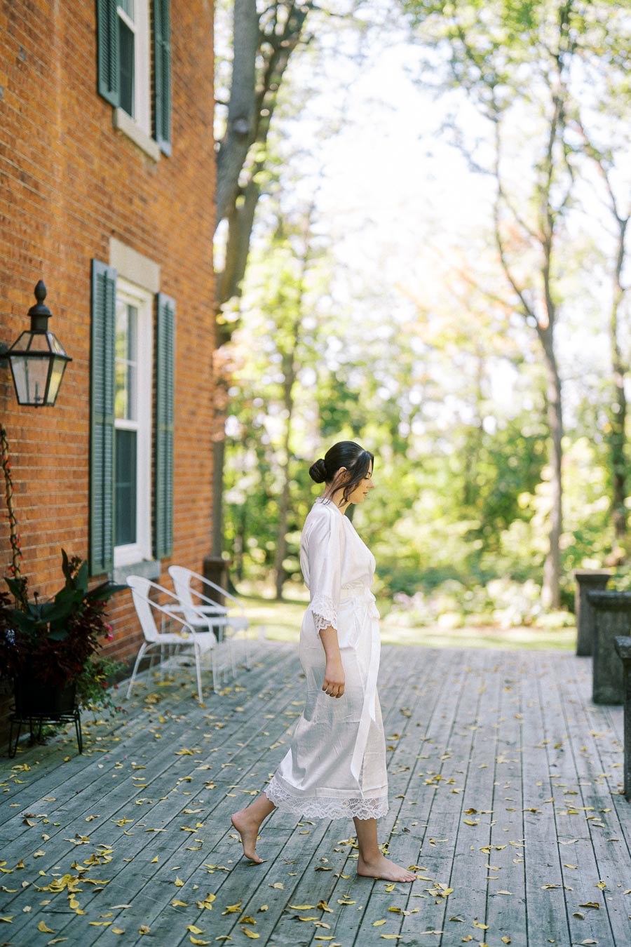 A woman in a white robe walks barefoot on a wooden deck surrounded by a lush garden, next to a brick building with green shutters.