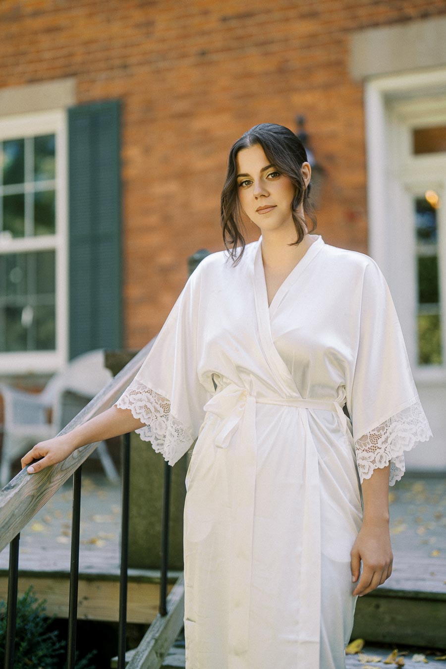 A woman in a white robe with lace details standing outdoors in front of a brick building, leaning on a wooden railing.