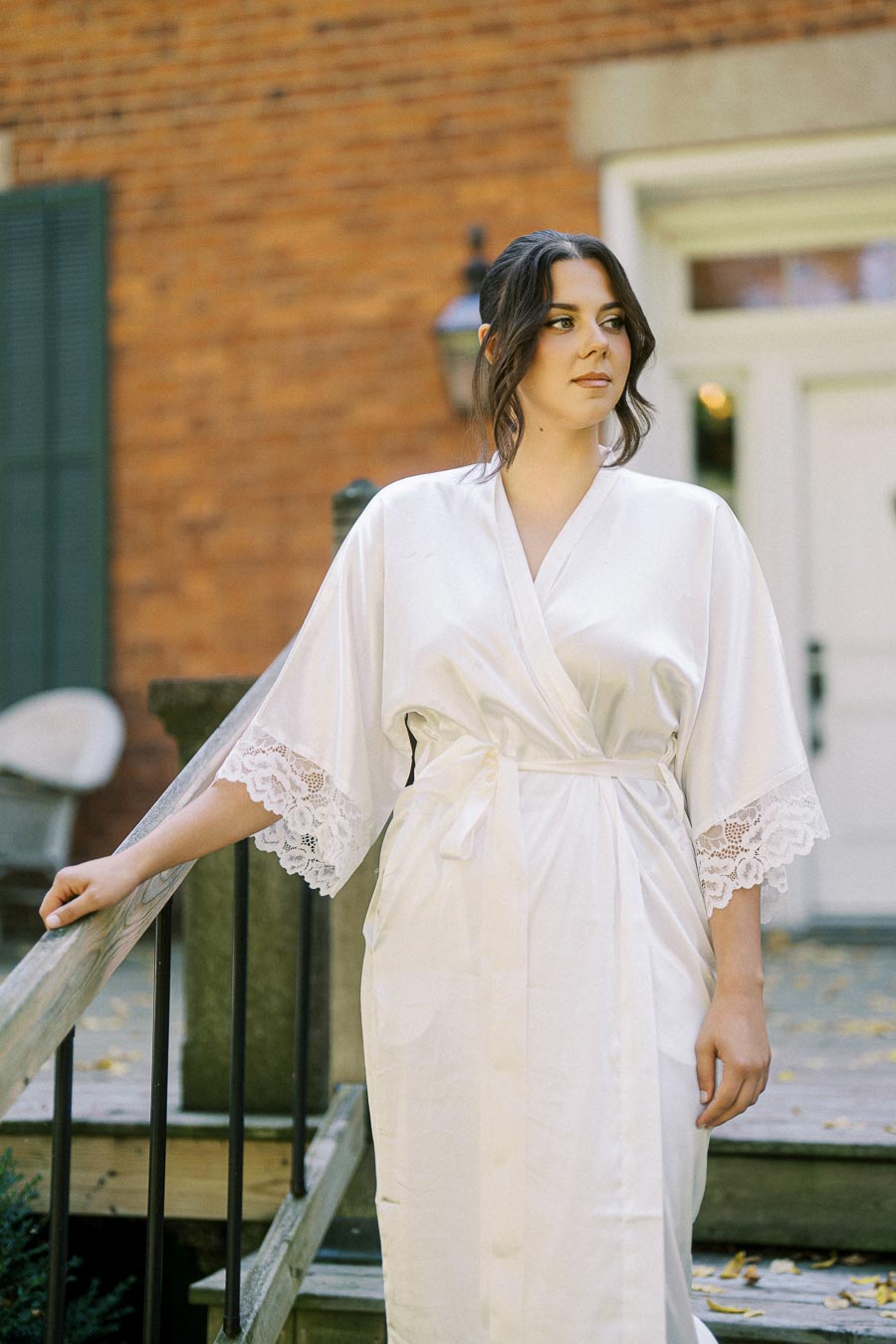 Woman wearing a white silk robe with lace sleeves, standing outdoors on a wooden staircase, with a brick building in the background.