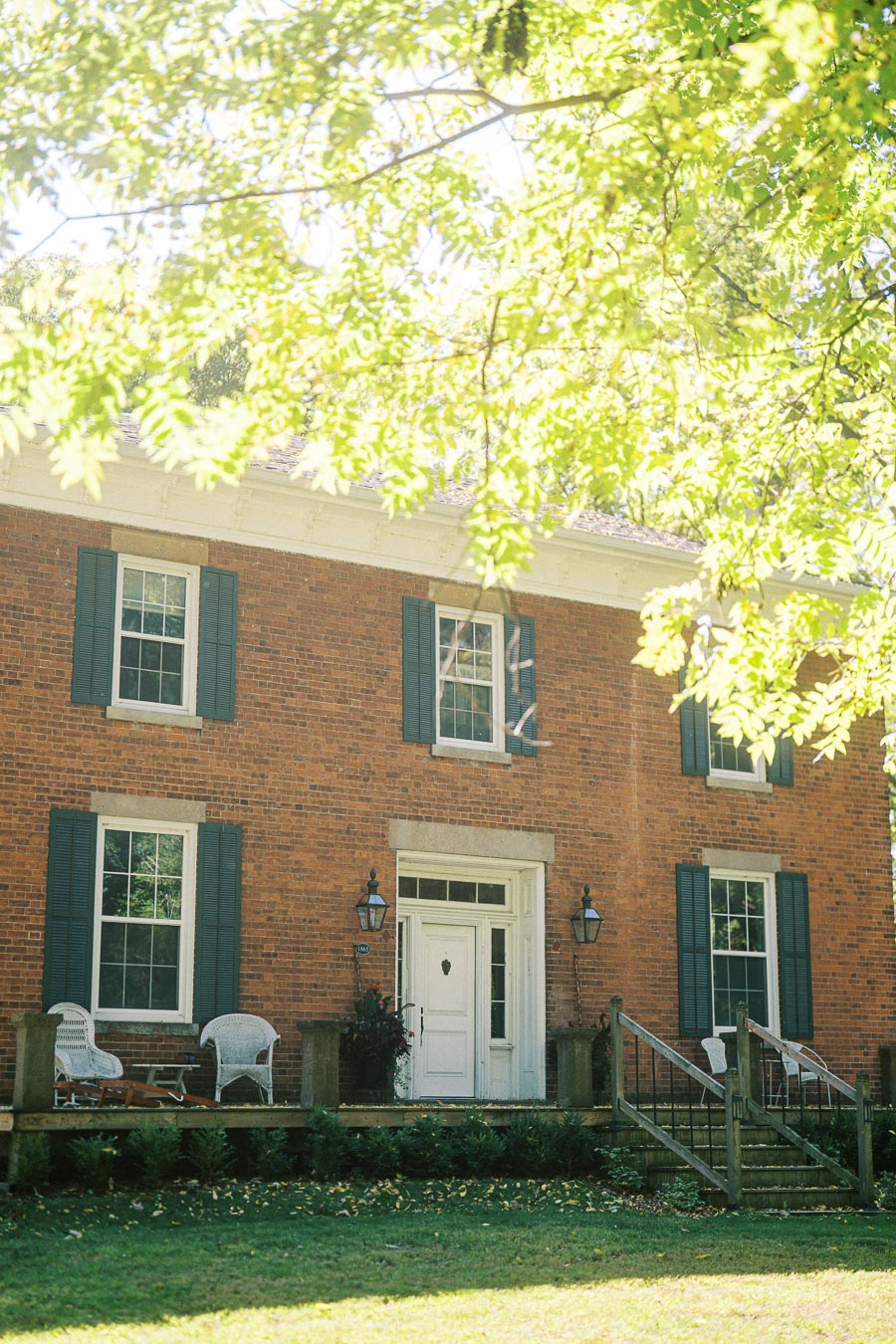 A charming, historic brick house with blue shutters and a white front door, surrounded by lush green trees and a well-maintained lawn basking in sunlight.