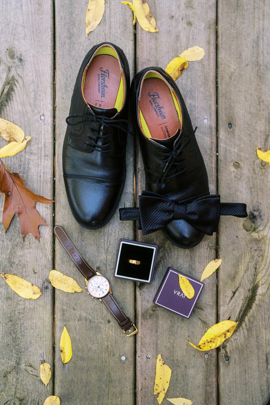 Stylish men's wedding accessories including black leather shoes, a matching bow tie, a gold ring in a box, and a classic watch displayed on a rustic wooden deck among fallen autumn leaves.