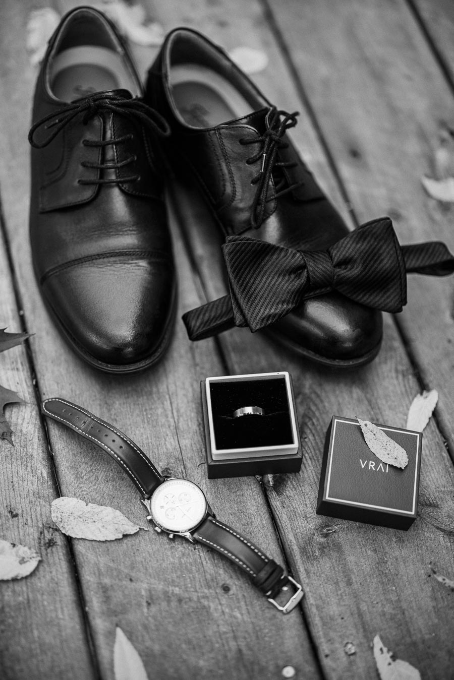 Black and white image of groom accessories on wooden surface, including black leather shoes, a black bow tie, a wristwatch, and a wedding ring in a box. Autumn leaves are scattered around, enhancing the elegant, rustic setting.