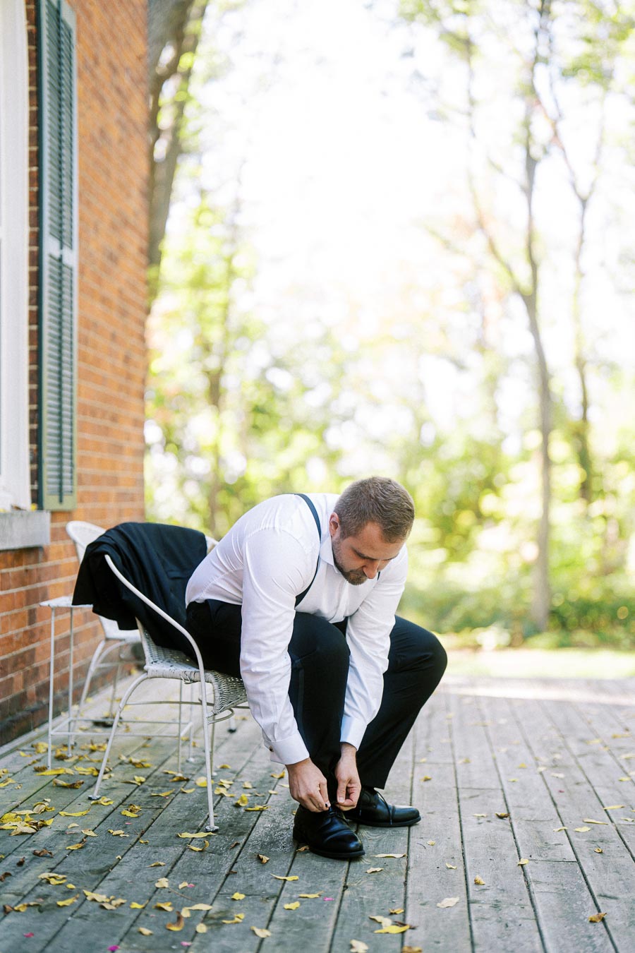Man tying his shoelaces on a wooden deck outdoors, wearing a white shirt and suspenders, jacket draped over a chair, with autumn leaves scattered on the ground.