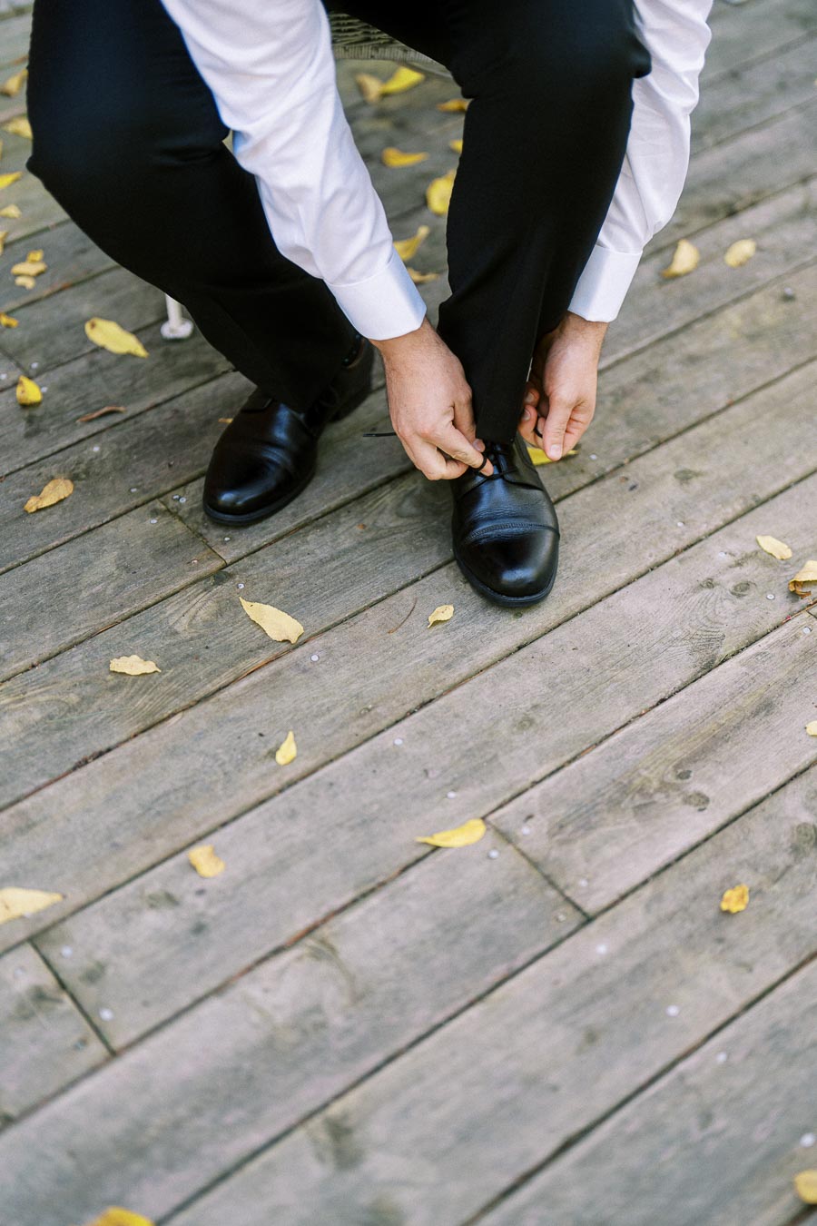 Man tying black dress shoes on wooden deck with scattered yellow leaves.