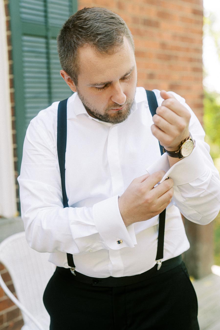 A man in a white dress shirt with suspenders adjusts his cufflinks outdoors, preparing for a formal event.
