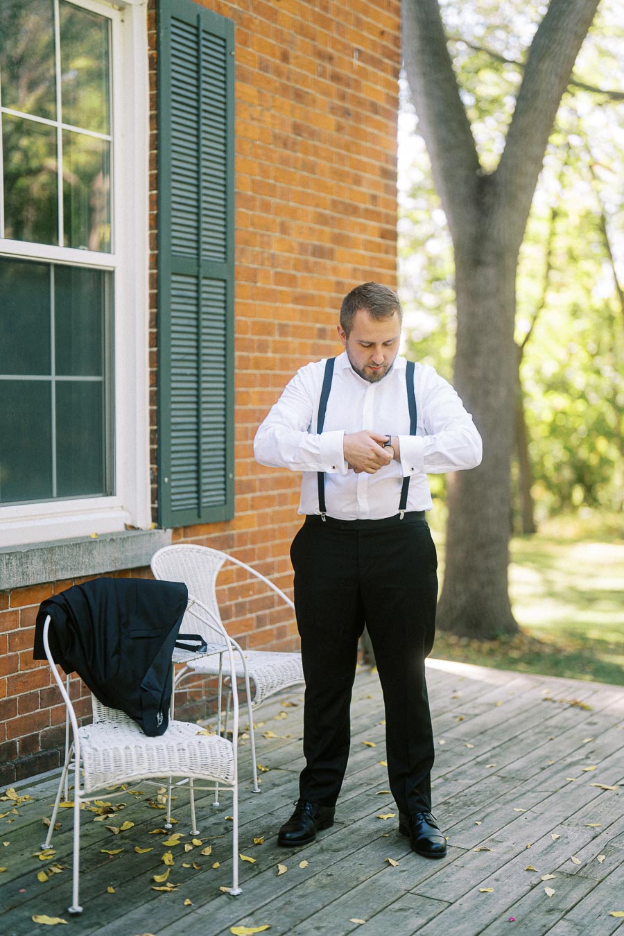 Man adjusting watch outdoors on a sunny day, wearing a white shirt with suspenders and black trousers, standing near a brick building and patio chair with a jacket.