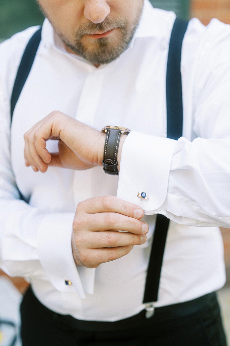 Man adjusting cufflinks on white dress shirt with suspenders and wristwatch.
