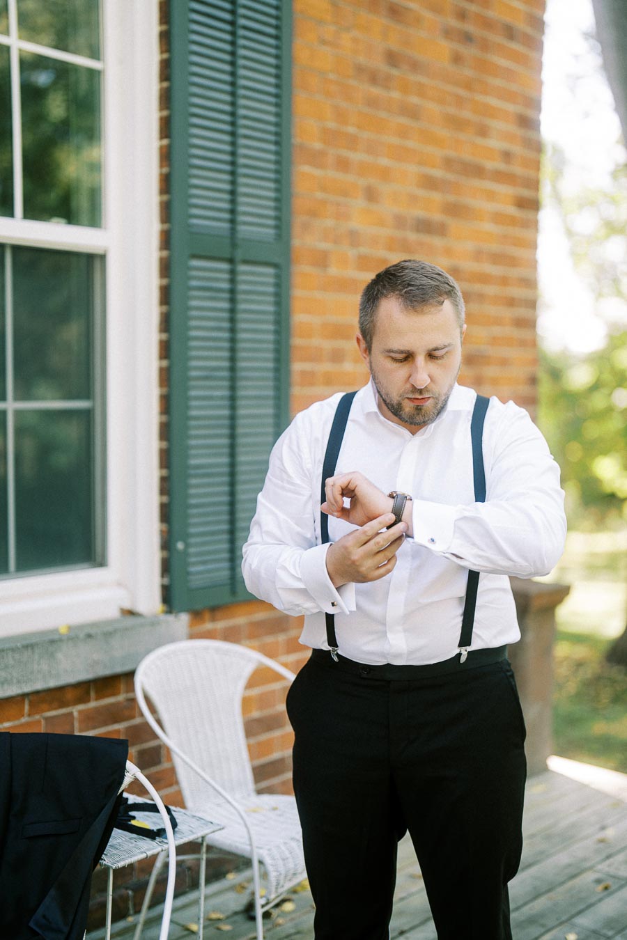 A man in formal attire with suspenders adjusts his wristwatch while standing on a wooden porch in front of a brick building with green shutters.