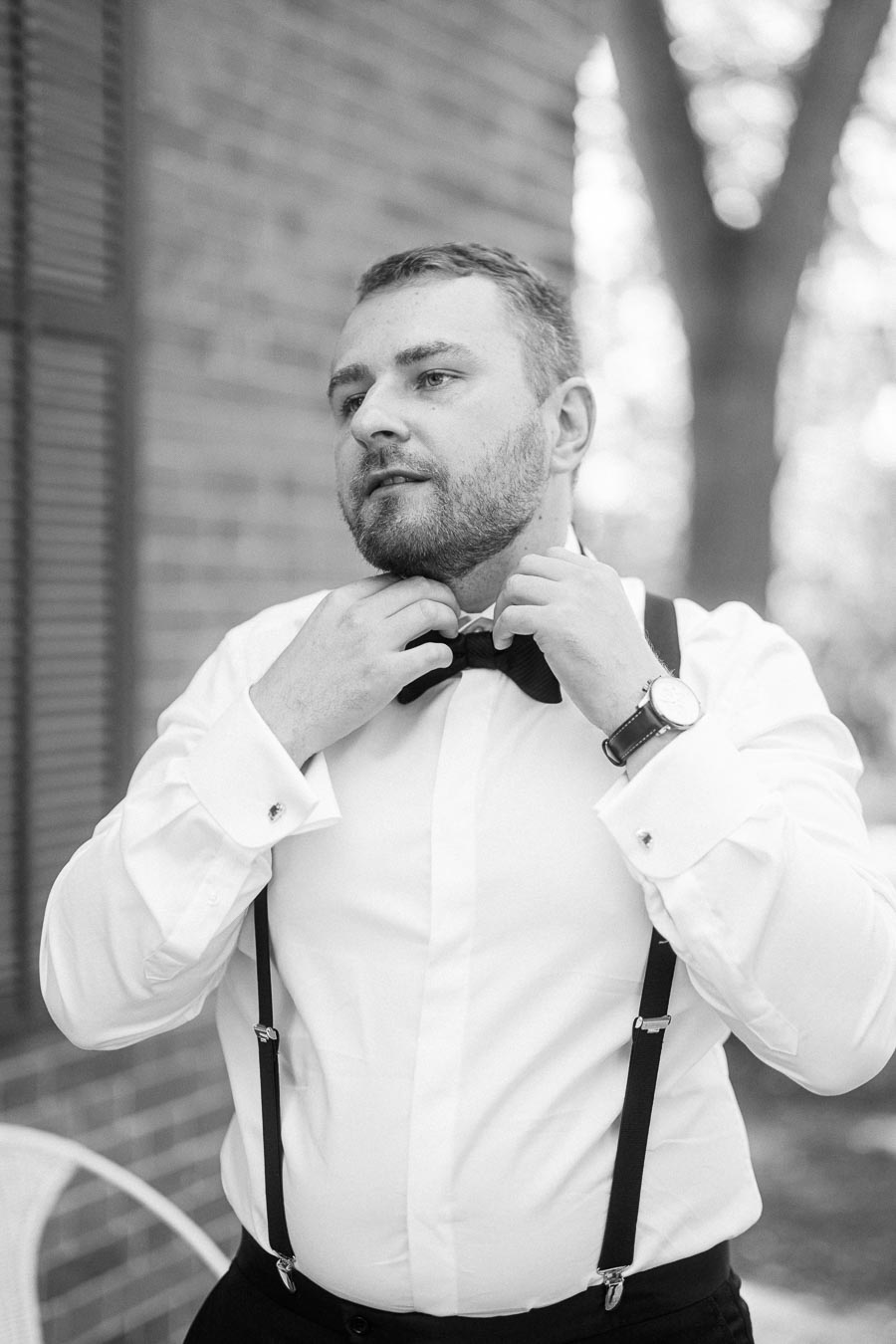 Black and white photo of a man adjusting a bow tie, wearing a formal white shirt with suspenders and a wristwatch, standing outdoors with a building and trees in the background.