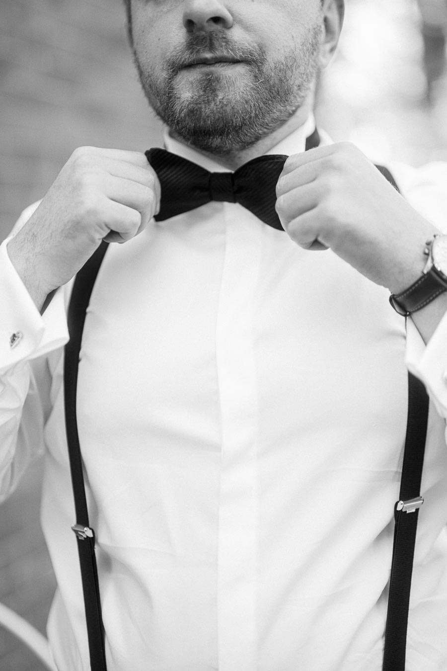 A man adjusts his black bow tie while wearing a white dress shirt with suspenders, exuding elegance and sophistication in a close-up black and white photograph.