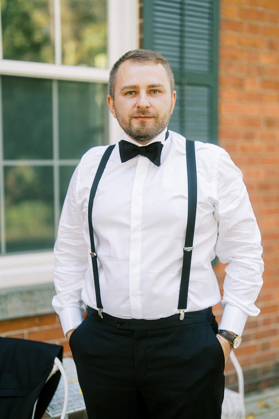 A man in a formal white shirt with black suspenders and a bow tie poses outdoors in front of a brick building with a window and green shutters.