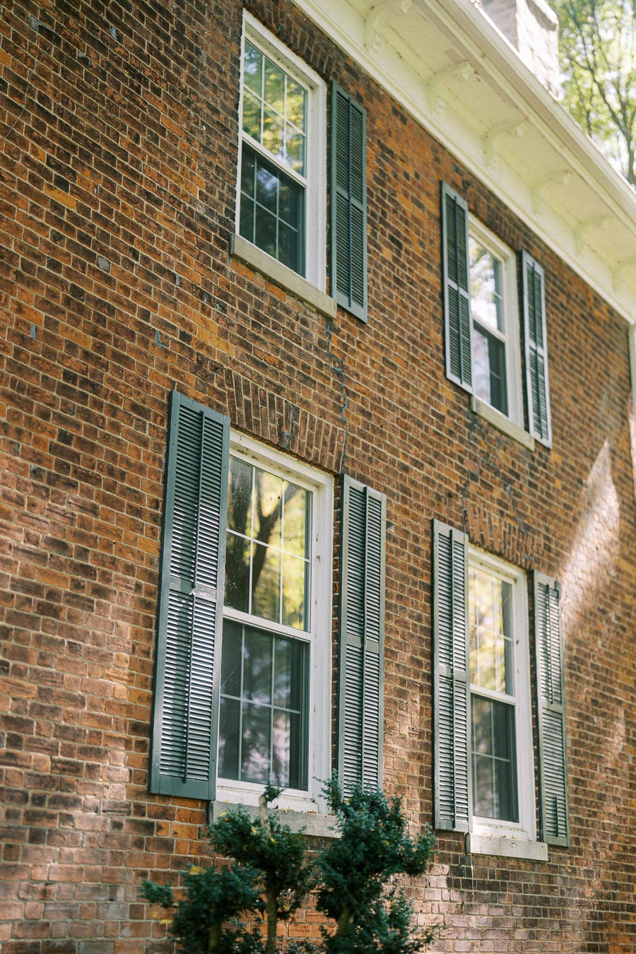 Exterior of a traditional brick house with double-hung windows, white trim, and green shutters, surrounded by lush greenery on a sunny day.