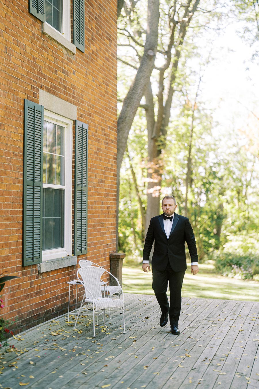 A man in a black tuxedo walks on a wooden deck beside a brick house with green shutters, surrounded by lush trees and greenery on a sunny day.