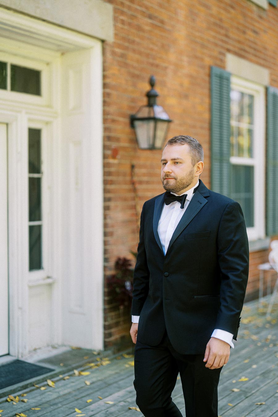 A man in a black suit with a bow tie stands confidently outdoors in front of a brick building with white trim and a lantern. The setting exudes elegance and style, ideal for a formal event or wedding.