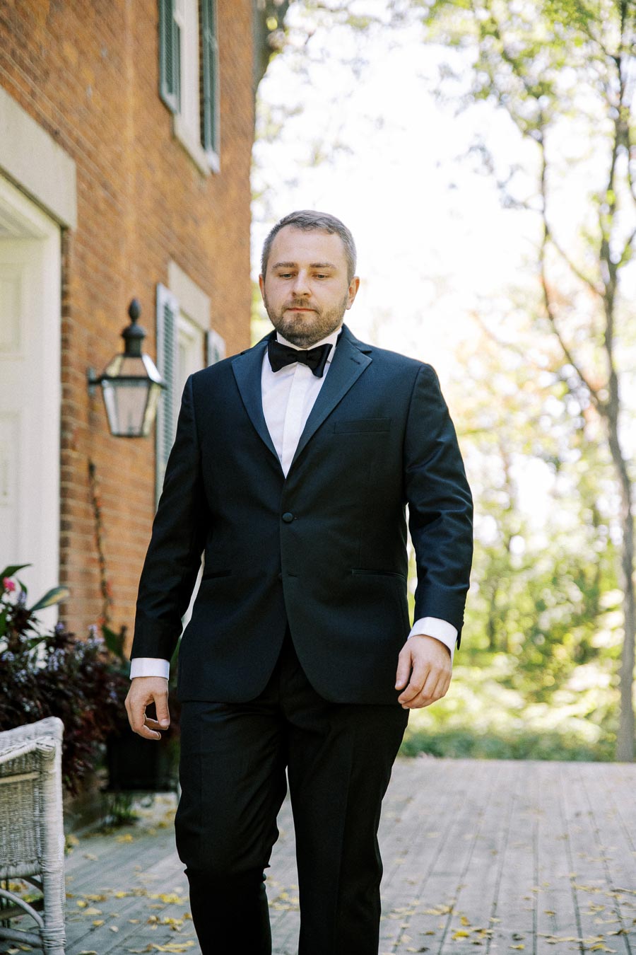 Man in a formal black suit and bow tie walking in front of a brick building with greenery in the background.