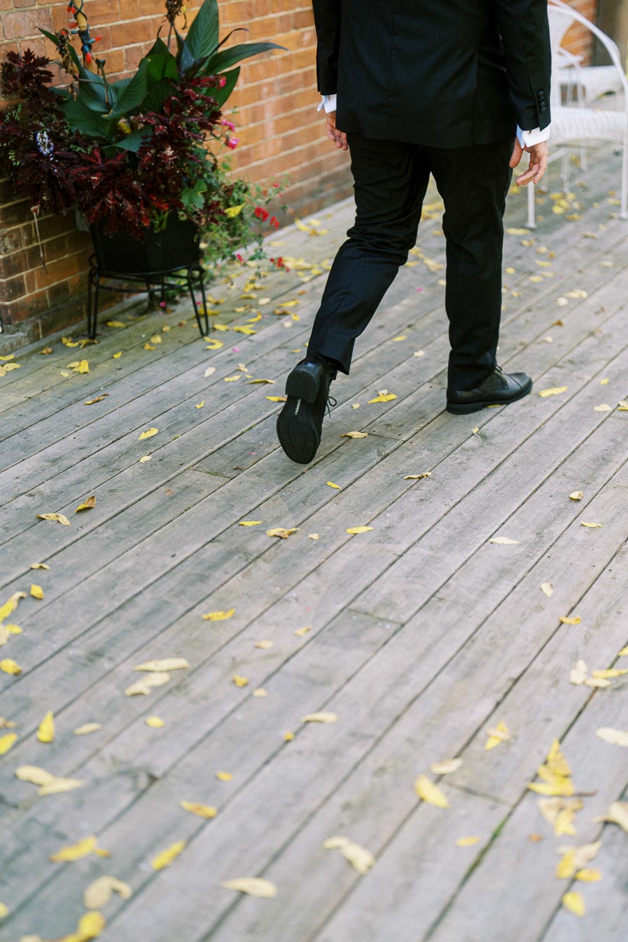 A person in a formal black suit walking on a wooden deck with scattered yellow leaves and a brick wall planter in the background.