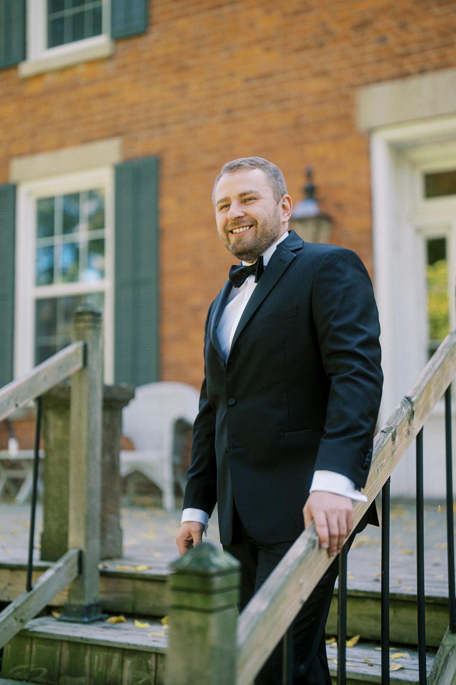 A man in a black tuxedo smiling while standing on a wooden staircase outside a brick building.