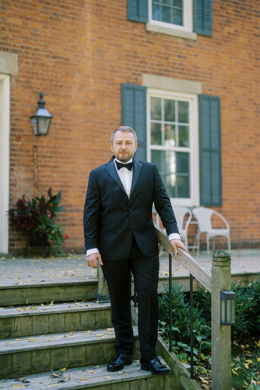 A man in a black tuxedo standing on wooden steps in front of a brick building with blue shutters, holding the railing while looking at the camera.