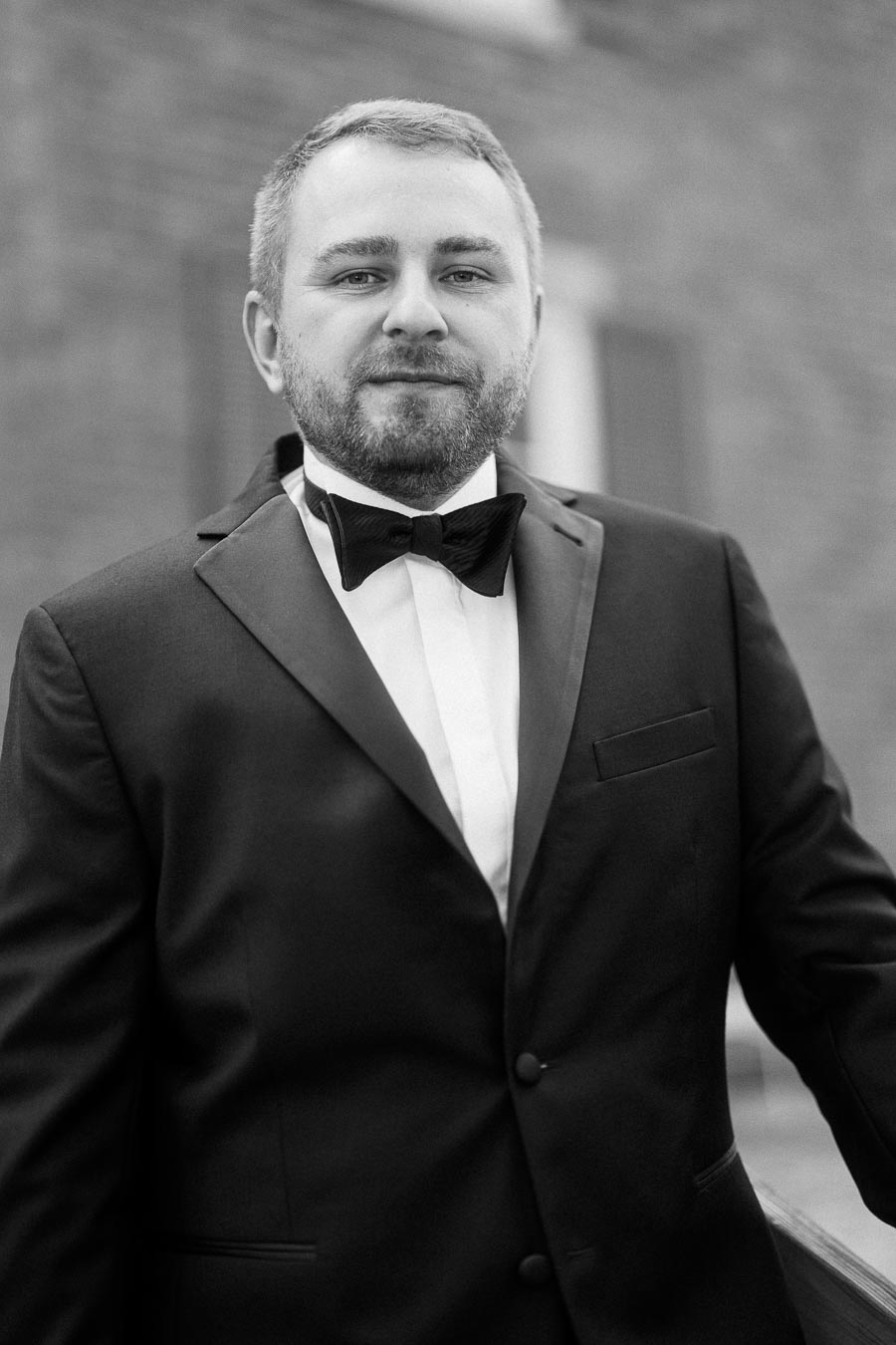 Formal portrait of a bearded man wearing a tuxedo and bowtie, standing against a blurred background, showcasing classic formalwear.