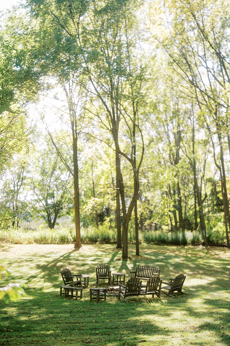 A serene outdoor scene featuring a circle of rustic wooden chairs arranged around a fire pit on a lush green lawn, surrounded by tall trees under a bright, sunny sky.