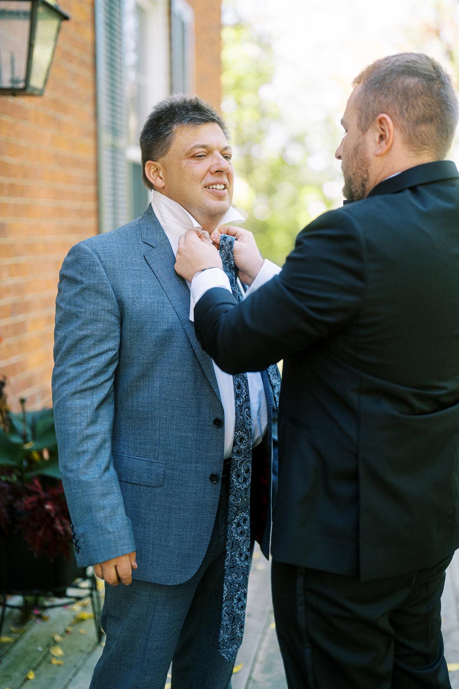Two men in suits on a sunny day, one adjusting the other's tie in front of a brick building.