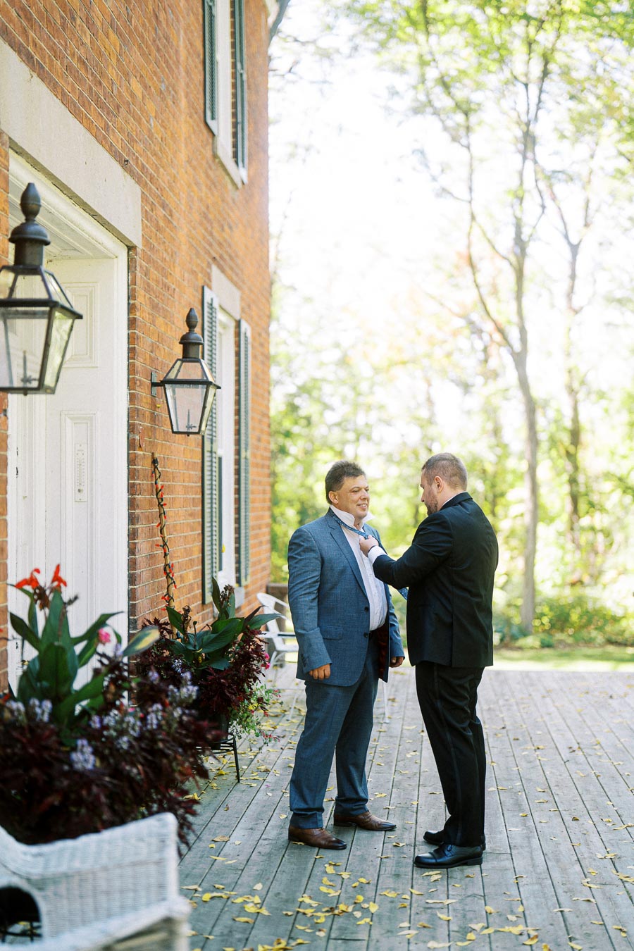Two men in suits adjusting a tie on a wooden deck outside a brick building, surrounded by greenery and decorative plants, with soft sunlight filtering through the trees.
