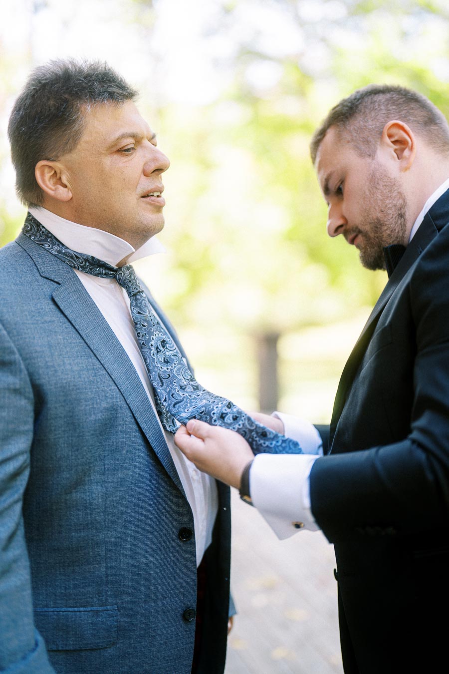 Two men in formal attire, with one adjusting the other's paisley tie outdoors.