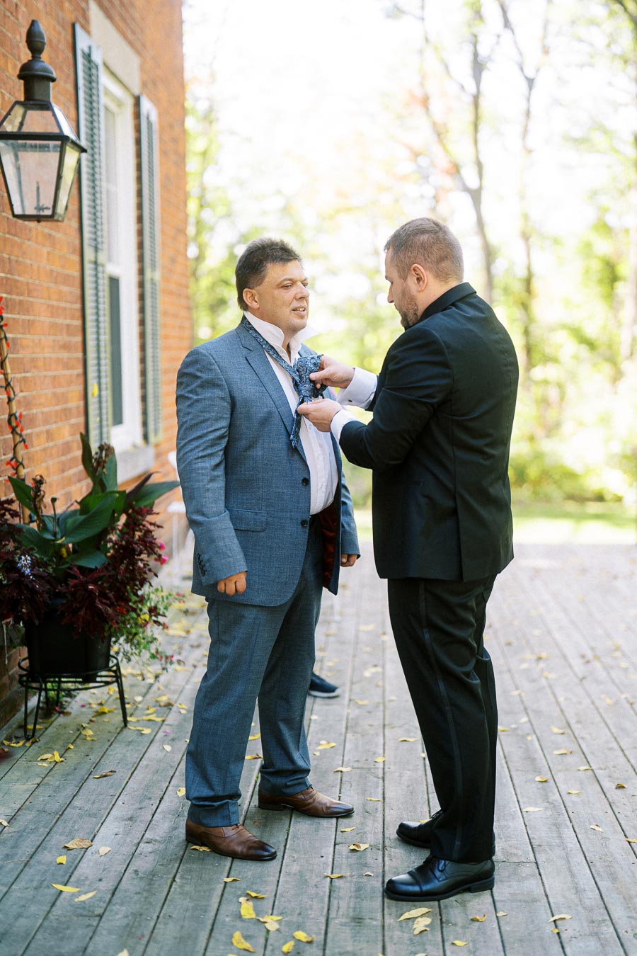 Two men getting ready for a formal event, with one adjusting the other's tie outside a brick building.