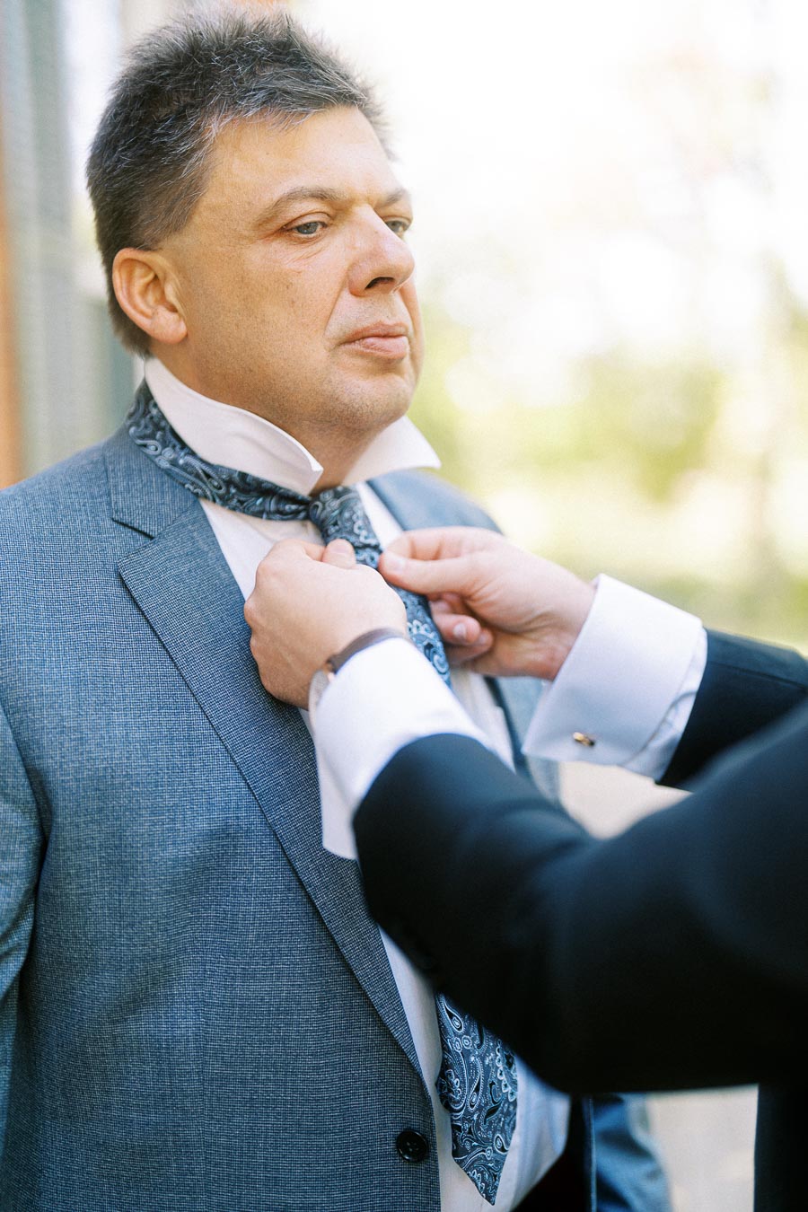 Man in a blue suit having his tie adjusted, outdoor setting with blurred background.