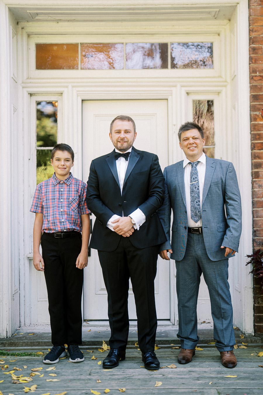 Three people posing in front of a white door; a young boy in a plaid shirt and dark pants, a man in a black tuxedo, and another man in a grey suit with a patterned tie, all smiling and standing on a wooden porch with scattered yellow leaves.