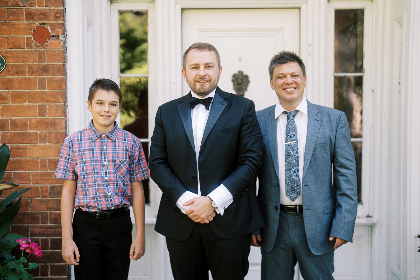 Three people posing in formal attire in front of a brick building with a white door. On the left, a young boy in a plaid shirt stands next to two men, one wearing a tuxedo and the other in a suit and tie, all smiling towards the camera.