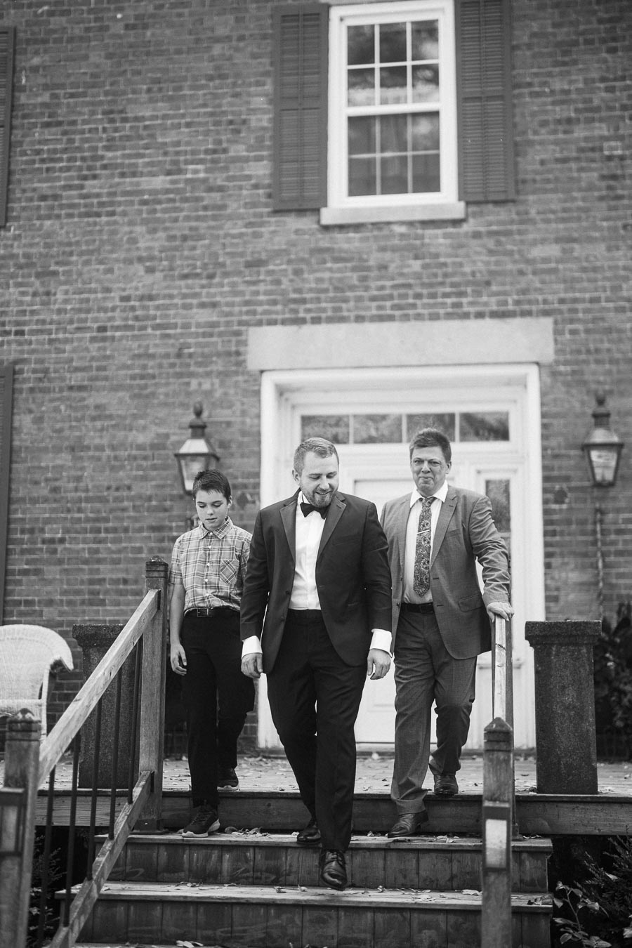Black and white image of three people dressed in formal attire descending stairs in front of a brick building with a window and shutters.
