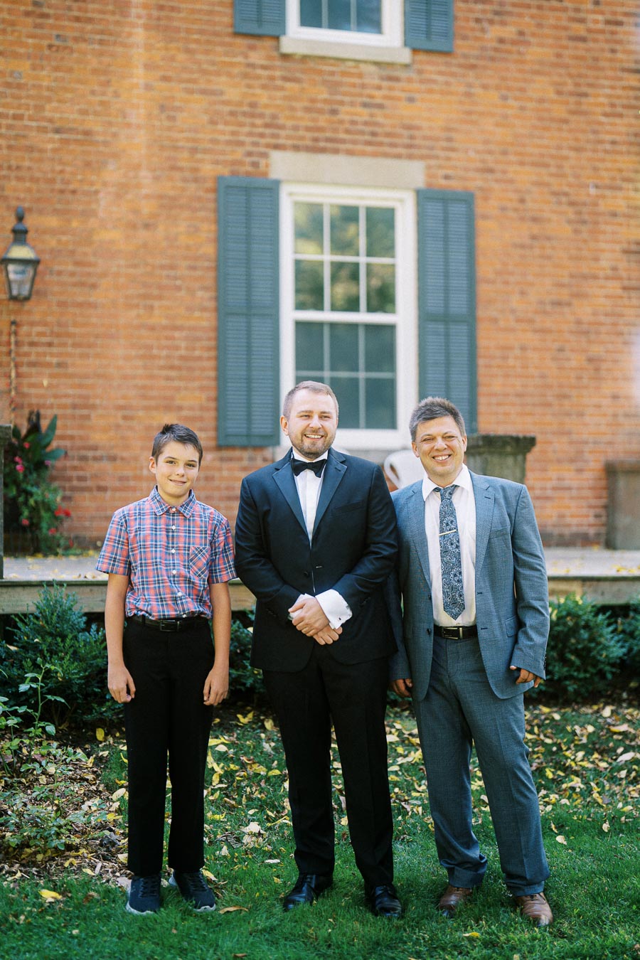 A group of three people smiling outdoors in front of a brick building with blue shutters and a window, wearing formal and casual attire, on a grassy lawn with greenery and fallen leaves.