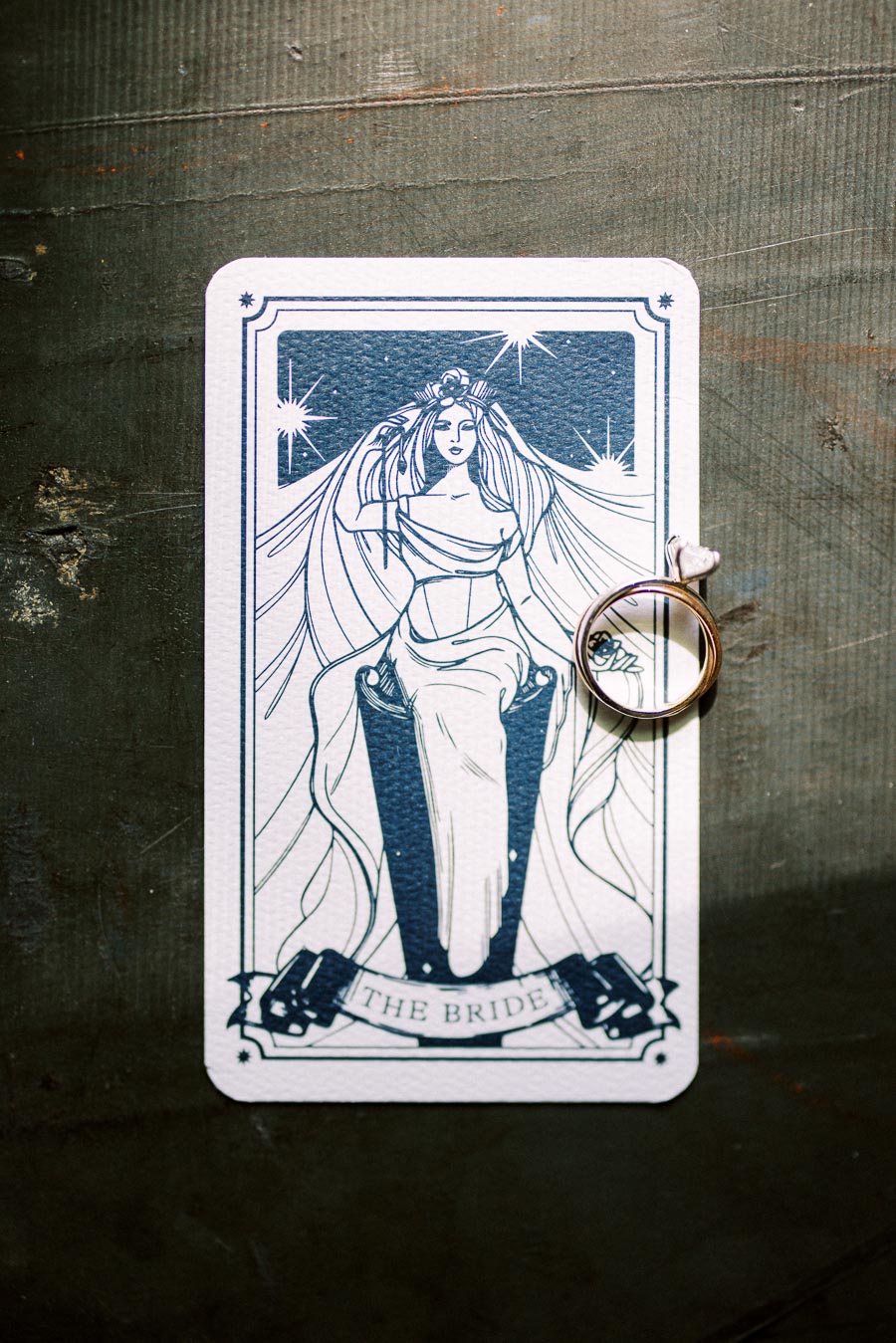 Tarot card labeled The Bride with a vintage-style illustration of a woman in a flowing gown, accompanied by a gold ring with a heart detail set on a textured gray surface.