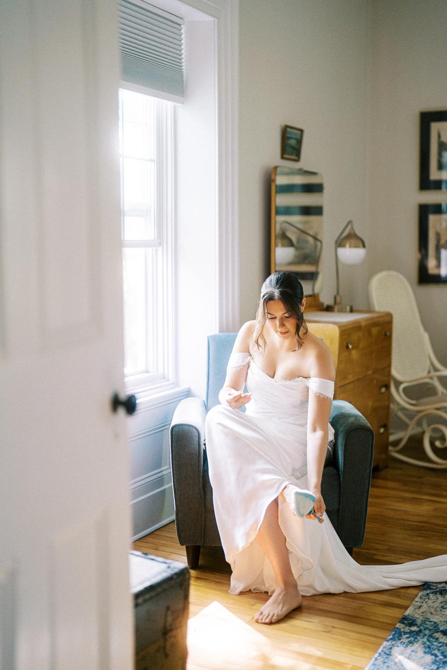 A bride sits on a chair in a softly lit room, putting on turquoise shoes, wearing an off-shoulder white wedding gown. The room features modern decor, including a wooden dresser, white walls, and a large window allowing natural light to fill the space.