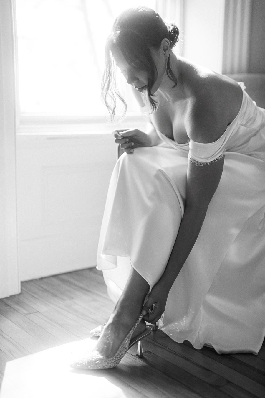 Black and white image of a bride in an elegant off-the-shoulder dress adjusting her sparkling high heels near a sunlit window, capturing a serene pre-wedding moment.