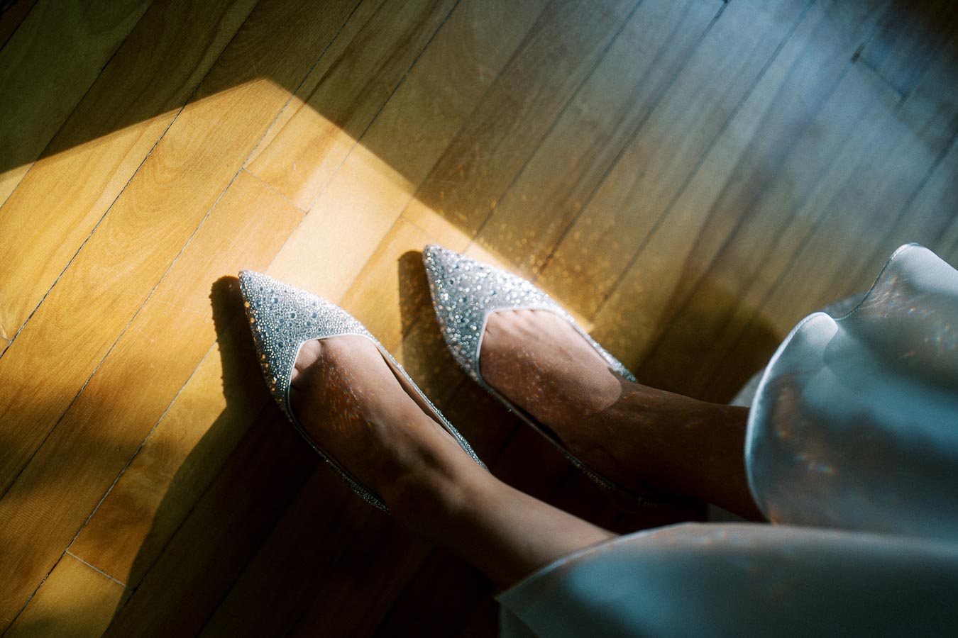 A close-up of a person wearing sparkly silver pointed-toe shoes on a wooden floor, with sunlight streaming through and creating a warm, elegant atmosphere.