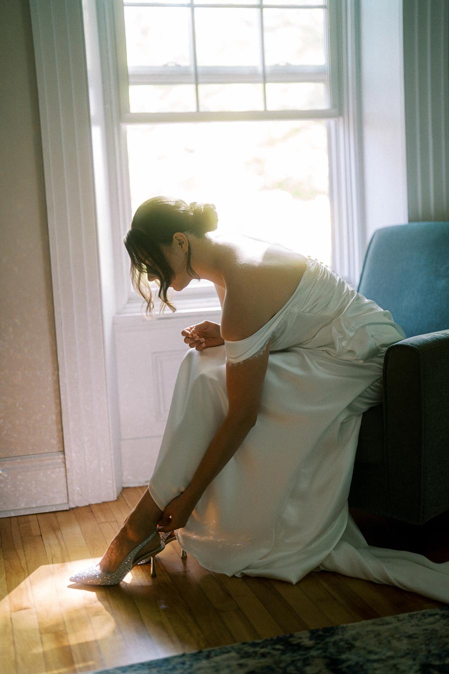 Bride adjusting her elegant sparkly heels in a sunlit room, wearing an off-shoulder white wedding gown beside a window and blue sofa.