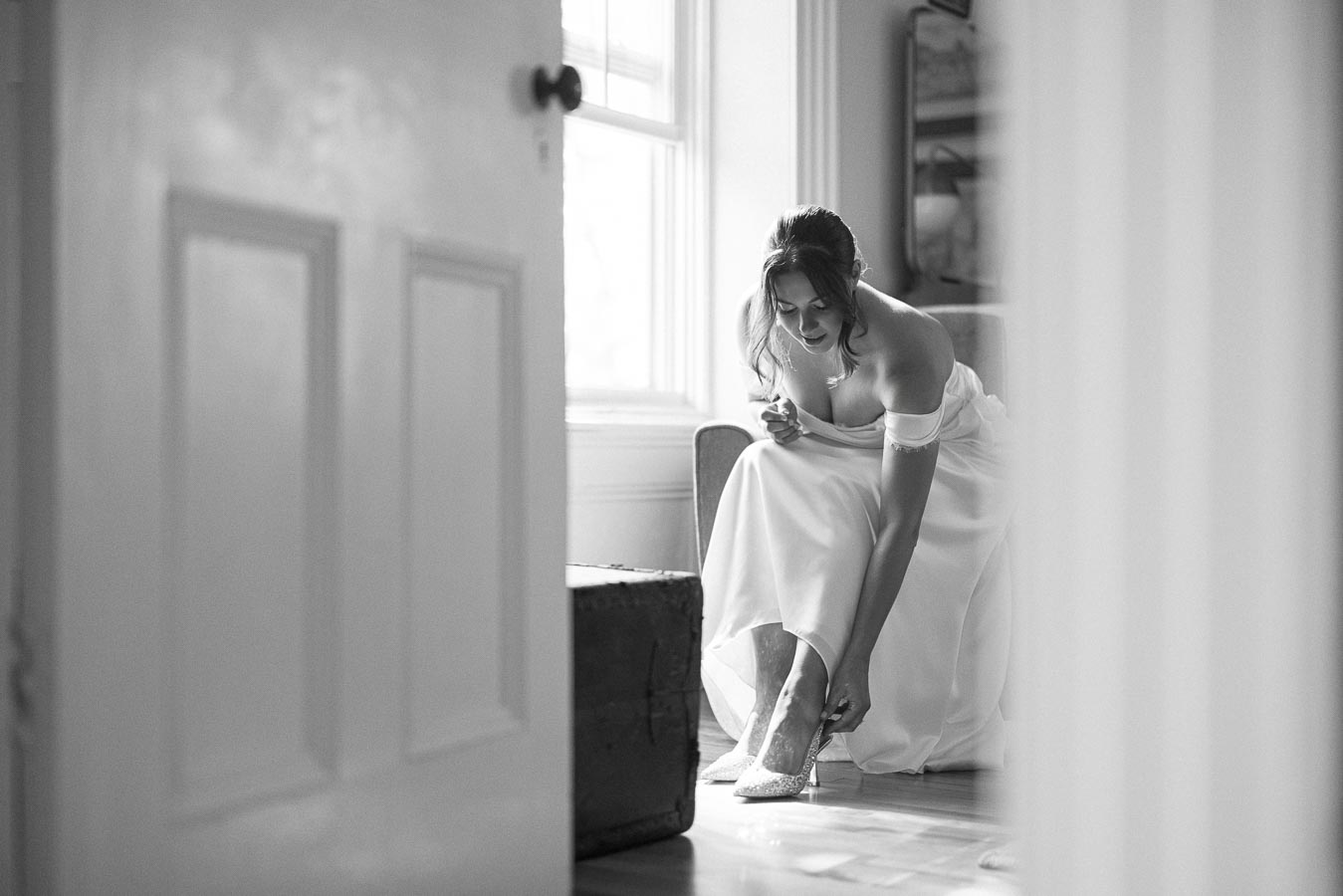 A bride in a white wedding dress, sitting near a window, adjusting her shoes, captured in black and white.