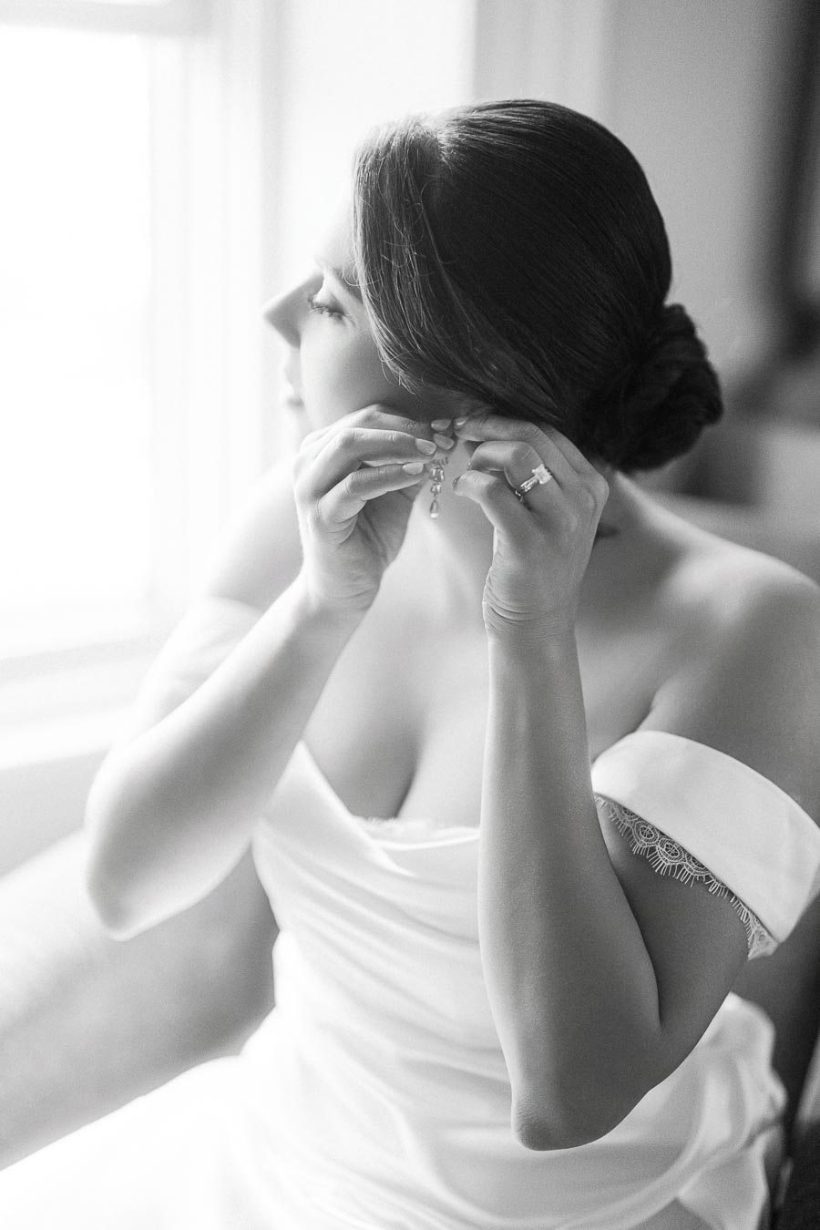 A bride in a white dress adjusting her earring while sitting by a window, showcasing elegant bridal style and jewelry in a soft, natural light setting.